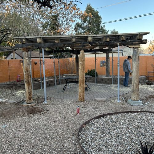 A rustic wood pergola under construction in a backyard patio in Albuquerque, New Mexico, featuring natural log posts, a slatted roof, and a paver stone floor surrounded by gravel landscaping.